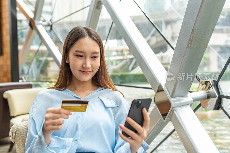 年轻聪明的亚洲女商人穿着蓝色寒意衬衫，正在通过智能手机用她的信用卡在线支付购物