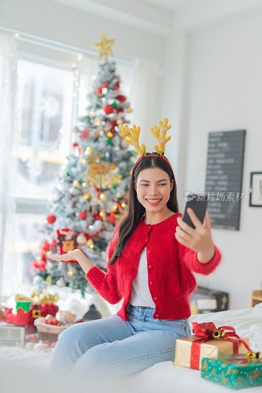 肖像照片的年轻美丽友好的亚洲女性与可爱的圣诞发带愉快地自拍与她的智能手机在她的房间一个漂亮的装饰圣诞树前