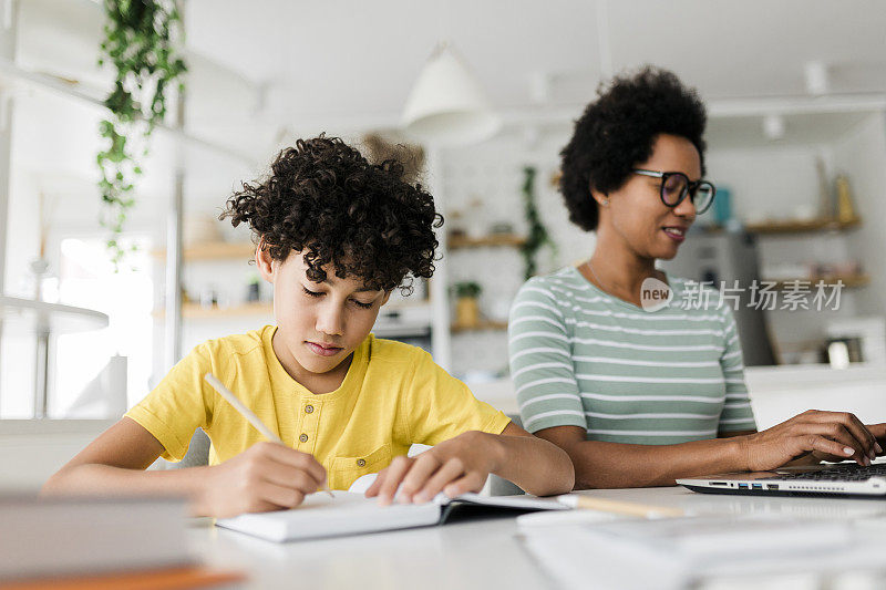 男孩在书上写字，而他的妈妈在笔记本电脑前工作。孩子在学习，母亲在家工作。
