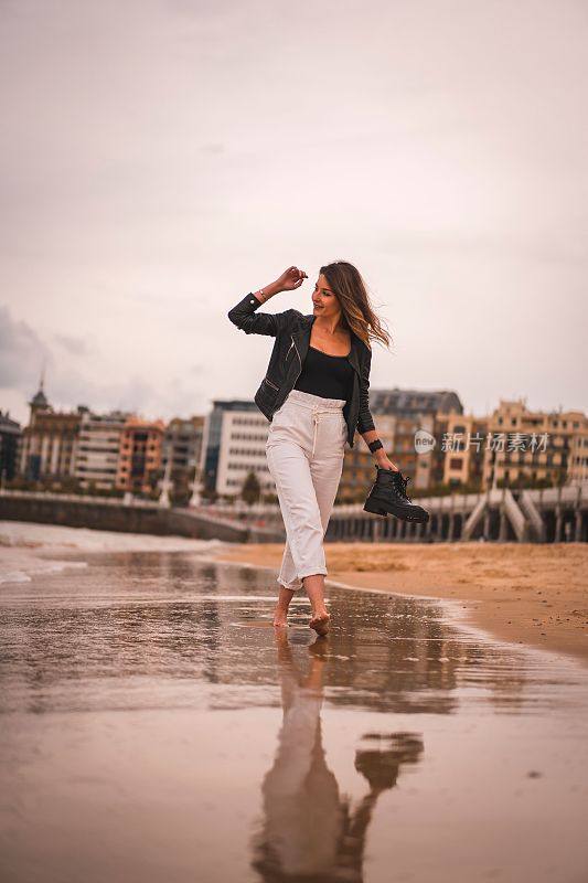 垂直拍摄的年轻时尚的女性在日落时赤脚走在海滩上，抱着她的海岸