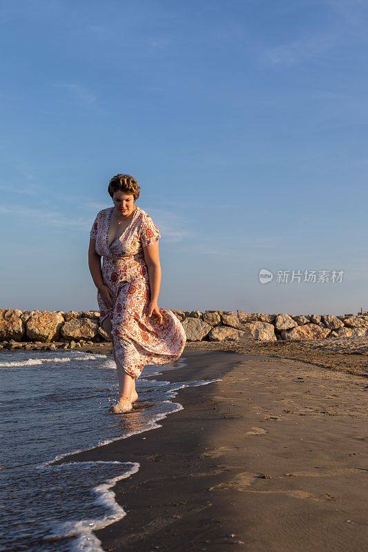 垂直拍摄一个曲线优美的白人女性在日落时走在海滩上