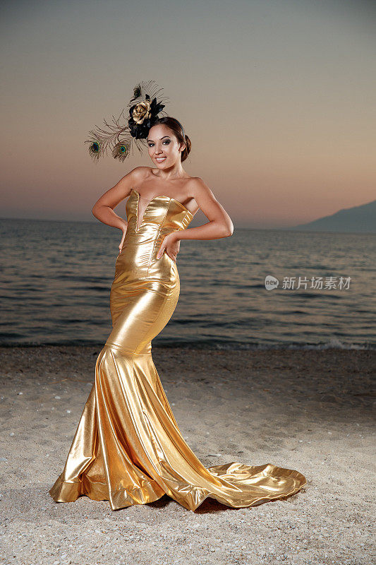 美丽的年轻女子在长金色无肩带连衣裙和羽毛头饰的头发，站在沙滩上在黄昏
