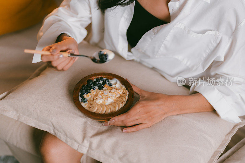 年轻的亚洲女性在家里客厅的沙发床上吃健康的早餐，有水果和酸奶