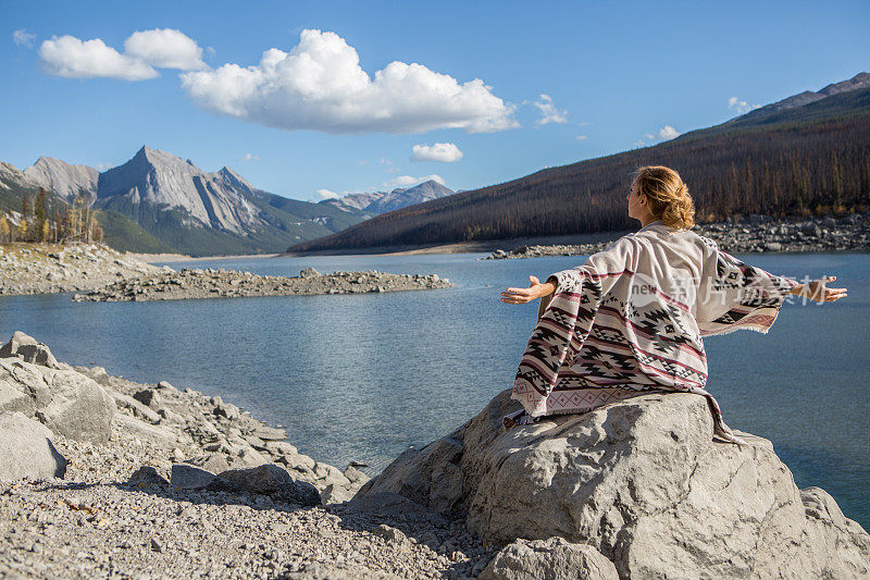 年轻女子坐在湖边的岩石上，胳膊伸着