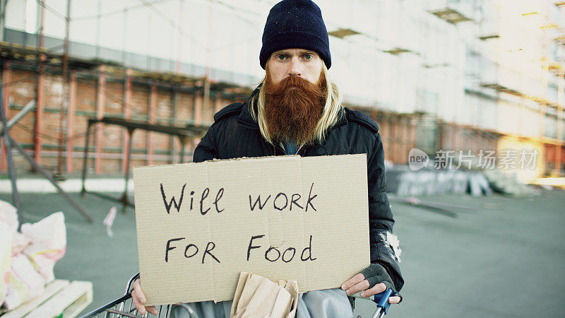 这是一个无家可归的年轻人的肖像，他带着硬纸板看着相机，想在购物车附近的食物站工作
