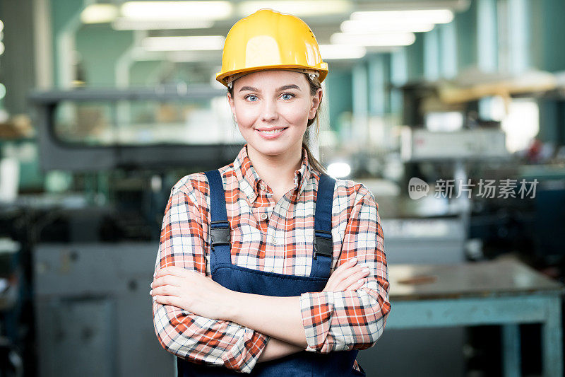 开朗漂亮的年轻女工人戴着黄色安全帽，在镜头前微笑，在工厂胸前交叉双臂
