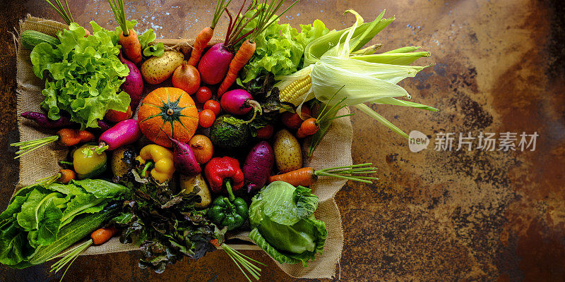 许多彩色的对比色沙拉蔬菜在麻袋材料坐在一个木制蔬菜托盘上，在一个抽象的陶土乡村背景与大气的照明。