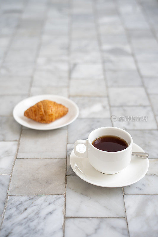 白杯子里装着牛角面包和黑咖啡，茶托放在大理石地板上