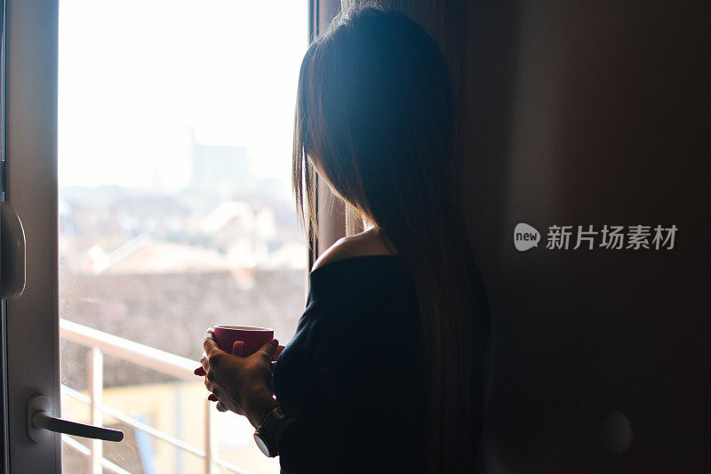 一个女人站在窗边，手里拿着咖啡杯。