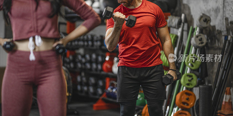 近距离的年轻运动男子是有哑铃的重量训练在健身房与年轻的女性健身教练的协助。