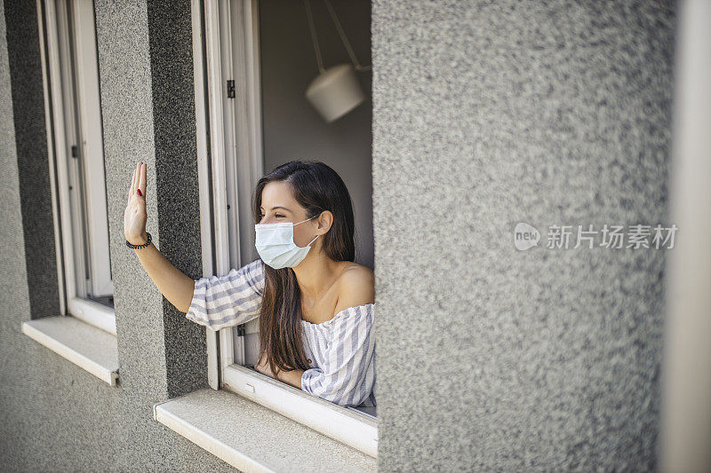 一名年轻的成年女子倚靠在一扇打开的窗户上，透过窗户向外看，并向邻居们挥手致意，而此时她正在家中休息，因为冠状病毒被隔离