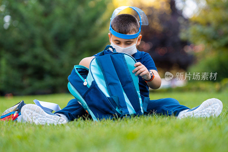 小男生戴着面罩和防护面罩，上学前坐在草地上检查书包