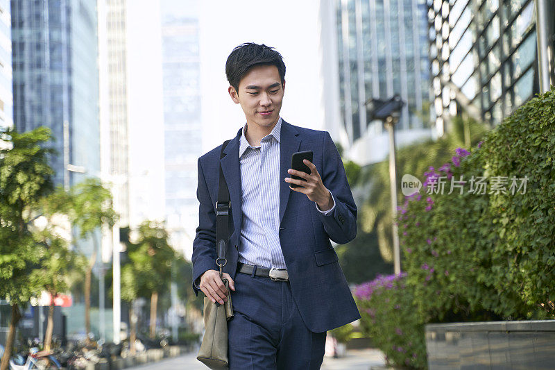 年轻的亚洲商人办公室职员一边走在街上一边看手机