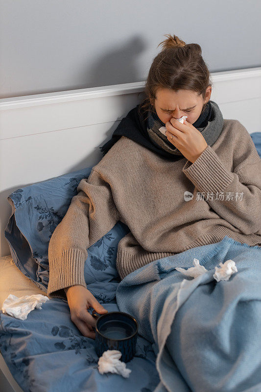 可爱的女人坐在床上，用纸巾擤鼻子，端着一杯热茶。女孩患有流鼻涕，鼻炎或鼻窦炎。季节性疾病。