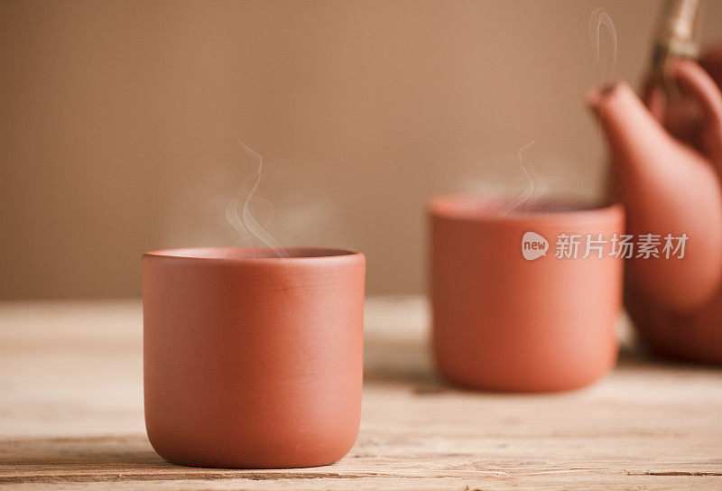 两个棕色陶瓷杯，茶或咖啡和热蒸汽，在质朴的木桌上，茶壶或咖啡壶的局部视图，棕色背景。空间的文本。
