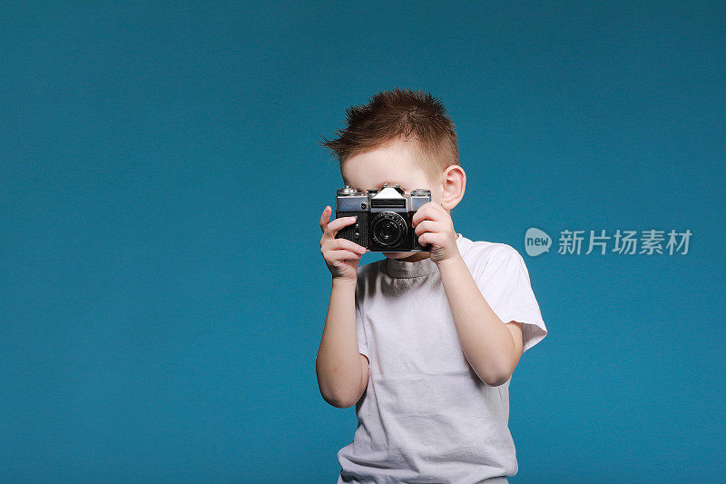 小男孩在用复古相机拍照。蓝色背景上的小男孩拿着老式相机。旧的技术概念和复制空间。孩子学习摄影