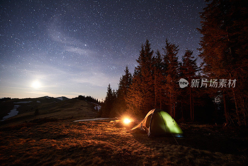 游客晚上在森林附近露营。在满是星星和月亮的美丽夜空下，照亮了帐篷和篝火