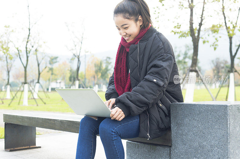 年轻女子坐在公园长椅上用笔记本电脑