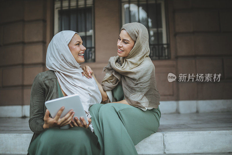 微笑的伊斯兰妇女戴着头巾，坐在楼梯上，手里拿着平板电脑