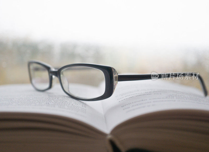 在雨天，眼镜放在一本打开的书上，背景是窗户