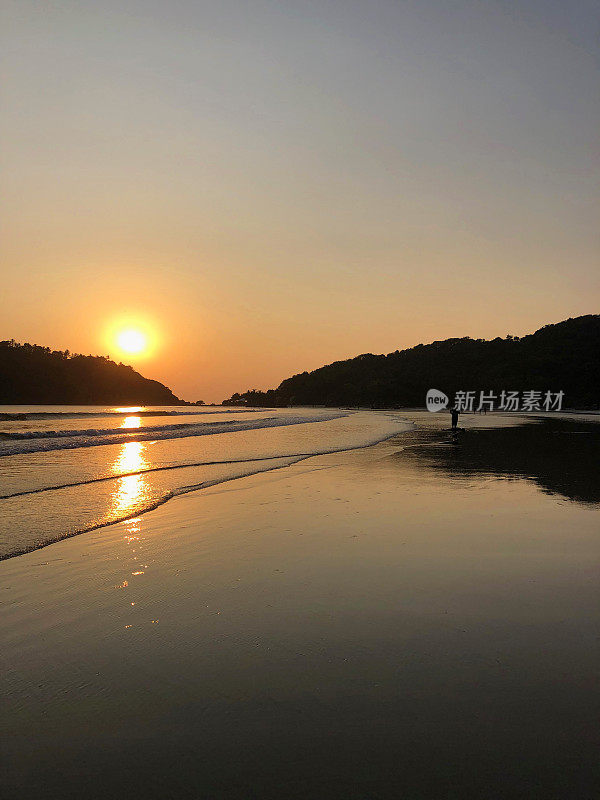 图片中，一名穿着短裤的男子走在印度果阿的帕洛伦海滩的海岸线上，夕阳下，海浪拍打着海滩