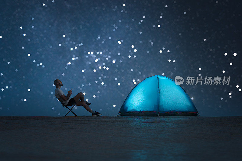一个男人坐在一个发光的帐篷旁边，看着夜空。手里拿着电话