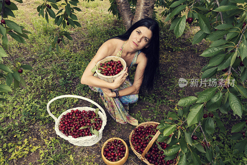 美丽的年轻女子穿着五颜六色的衣服，在樱桃树下，带着两个柳条篮子和两个装满水果的桶选择性的焦点