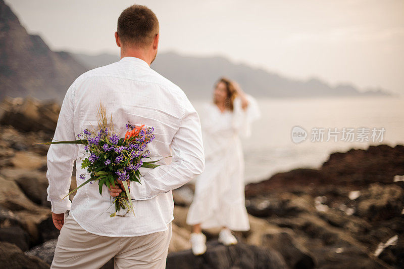 男新郎身穿白衬衫，背后捧着一束花站在海边看新娘