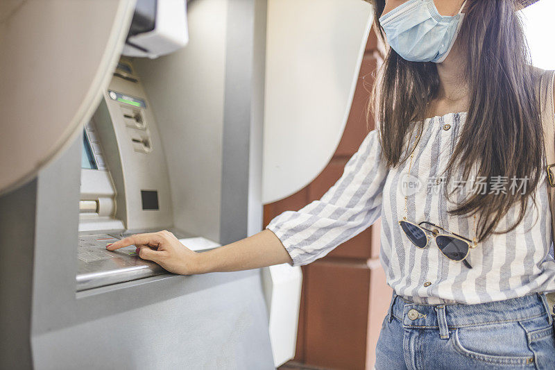 近距离的女人戴着保护面具站在城市街道上，使用ATM机取钱。冠状病毒或Covid-19病毒大流行概念。