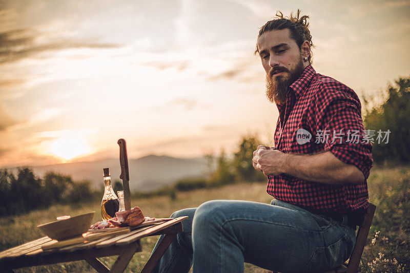 自然的守护者。一个在山上的男人，一个伐木工人，喝着自制的白兰地，吃着自制的食物，而在他身后是美丽的日落，休息辛苦的工作。