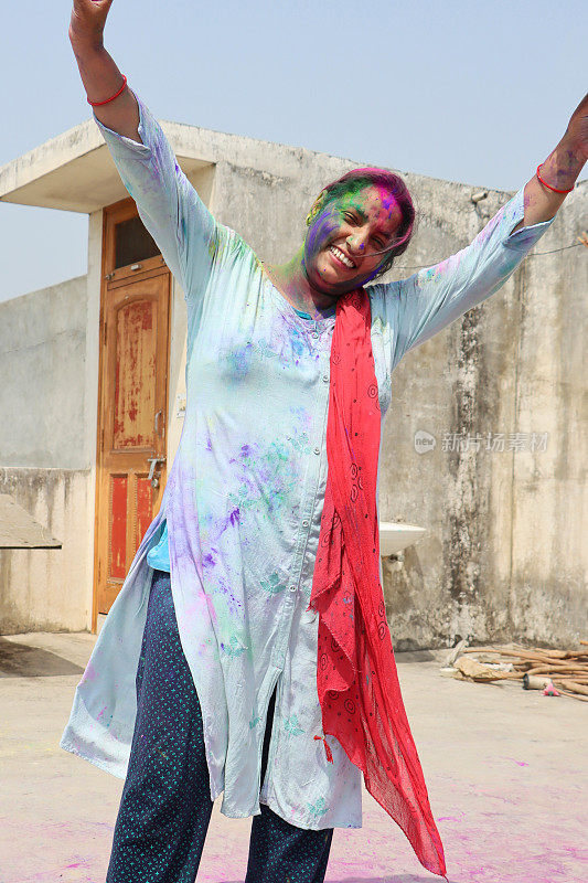 印度印度教妇女身穿沙尔瓦·克米兹的传统服装，站在涂满粉漆的地方举起双臂，胡里节的彩绘战斗，微笑着看着相机