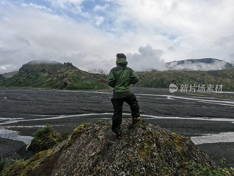 有一个人在冰岛徒步旅行，他站在一块岩石的顶端，眺望着山谷