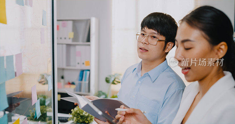 年轻的亚洲商人和女商人团队合作，穿着正装衬衫，手持电子平板电脑，站在透明的白板前头脑风暴会议和在工作场所写商业计划。