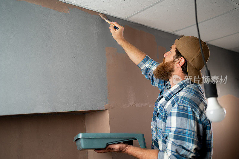 大胡子男人拿着画笔在灰色的墙上涂上橙色，留白为你的文字，复制空间，广告或设计。