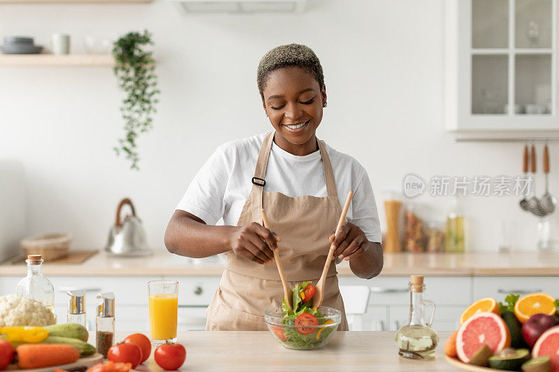 满意的千禧非洲裔美国女士在围裙准备有机蔬菜沙拉在餐桌上的现代厨房