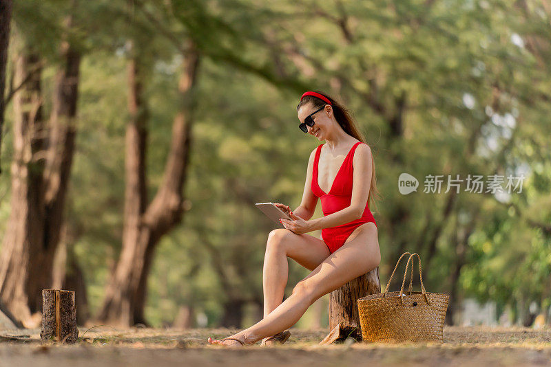 美丽的白人女性性感的女人在红色泳装比基尼与一个数字平板电脑，戴着太阳镜和一个红色的头带微笑，在海滩的树林里愉快地放松
