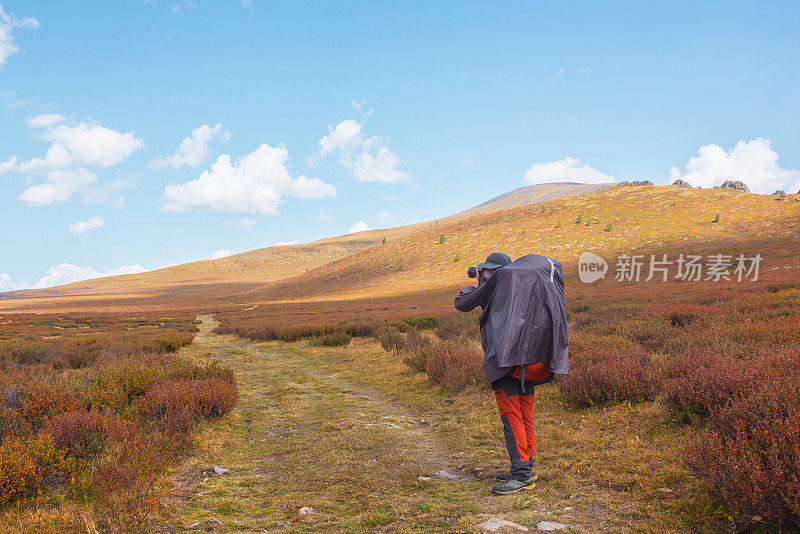 一个人穿着雨衣，背着大背包，在高山高原凋零的秋天植被中拍摄自然。背包客带着相机在多变的天气里拍摄山景