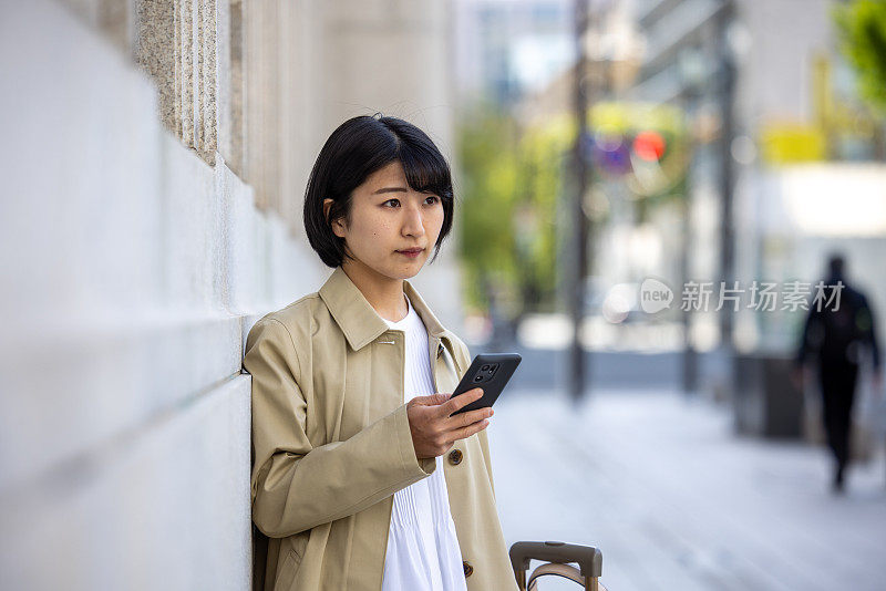 年轻的女商人靠在大楼的墙上，在城市里使用智能手机——等待