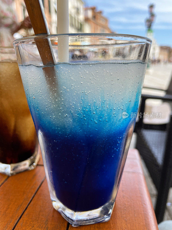 户外餐厅场景的特写图片，餐桌上摆放着蓝色覆盆子和可乐冰沙冰饮料，威尼斯海滨背景，重点放在前景