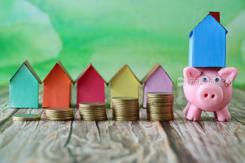 特写图像的一排多色纸板房子后面的一排排堆叠的硬币除了小猪银行平衡蓝色的模型房子背面，重点是前景，斑驳的绿色背景，家庭财务，房地产和假日储蓄的概念