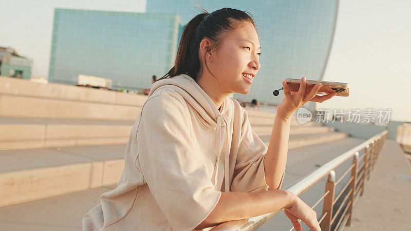 日出时，穿着运动服的亚洲女孩站在现代建筑背景的堤岸上发送语音信息