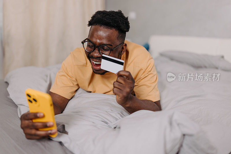 一个年轻人舒舒服服地躺在床上，手里拿着手机和信用卡，沉迷于网购