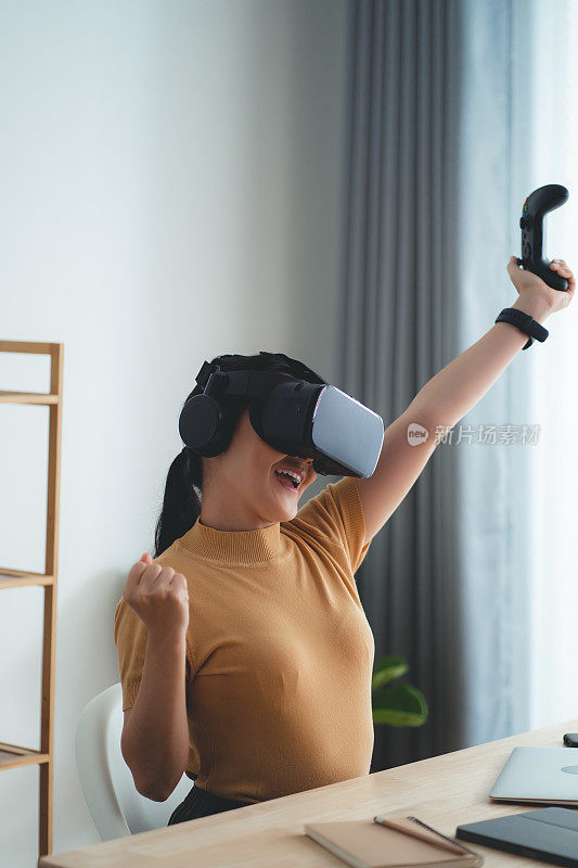 亚洲女性戴着VR头显，拿着操纵杆，在家庭办公室开心地微笑着。