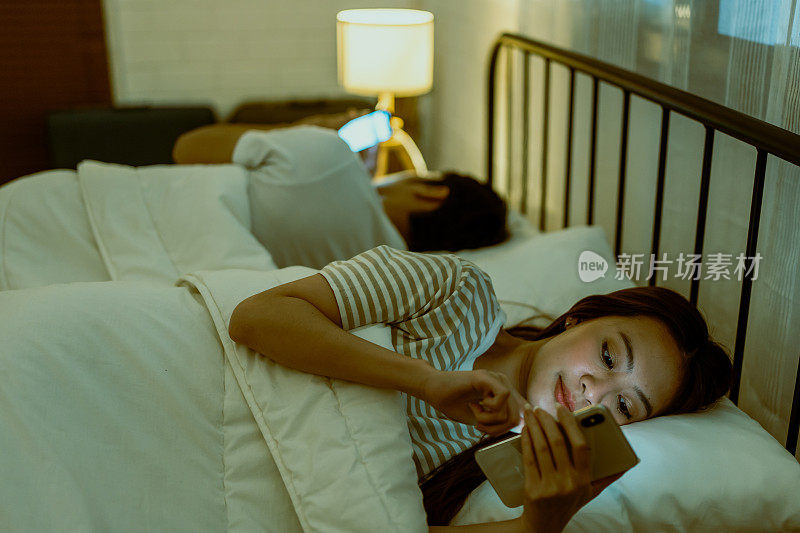 亚洲年轻的手机成瘾夫妇不关注彼此。家庭问题，新婚夫妻躺在床上玩智能手机，忽略了家里卧室里的夫妻。