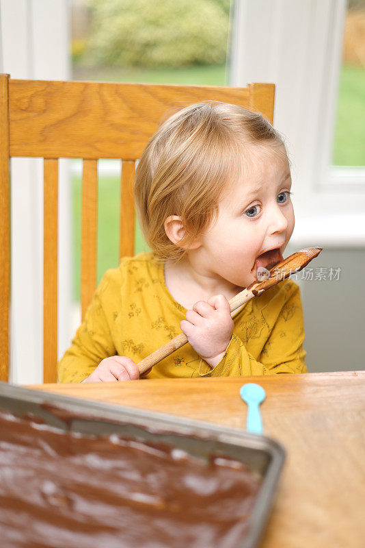 可爱的3岁小女孩，在厨房的桌子上做了她的第一个巧克力纸杯蛋糕，用木勺舔着剩下的混合物