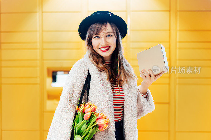 在现代邮政自动邮件终端机旁，一位面带微笑的时尚女性手持盒子的肖像，该终端机设有自动取件或退款装置。用于存放包裹的电子储物柜。有选择性的重点