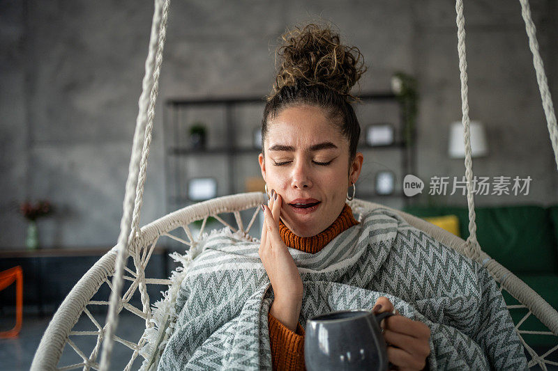 年轻女子喝着热茶或咖啡，感到牙痛，坐在家里客厅的秋千上。