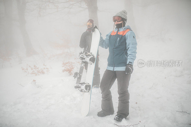 在一个雾蒙蒙的冬日，穿着全套冬季装备的男女滑雪板运动员站在山上，准备滑雪