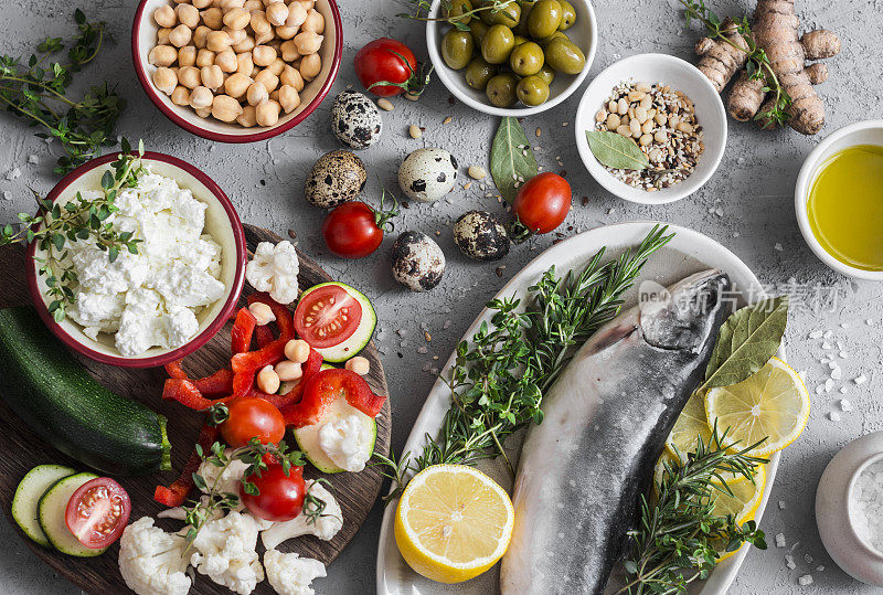 地中海风格的食物背景。鱼，蔬菜，香草，鹰嘴豆，橄榄，奶酪在灰色背景，俯视图。健康食品的概念。平躺