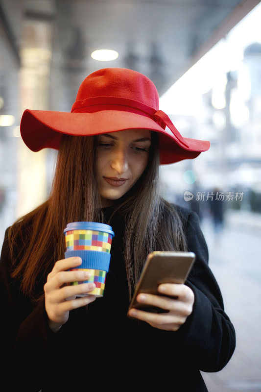 年轻女子戴着红帽子，在城市街道上使用智能手机和喝咖啡